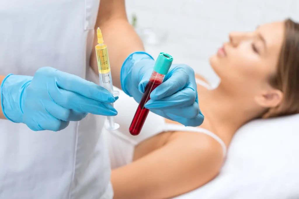 Gloved hands hold a syringe and blood-filled collection tube while a woman lies on a treatment bed.