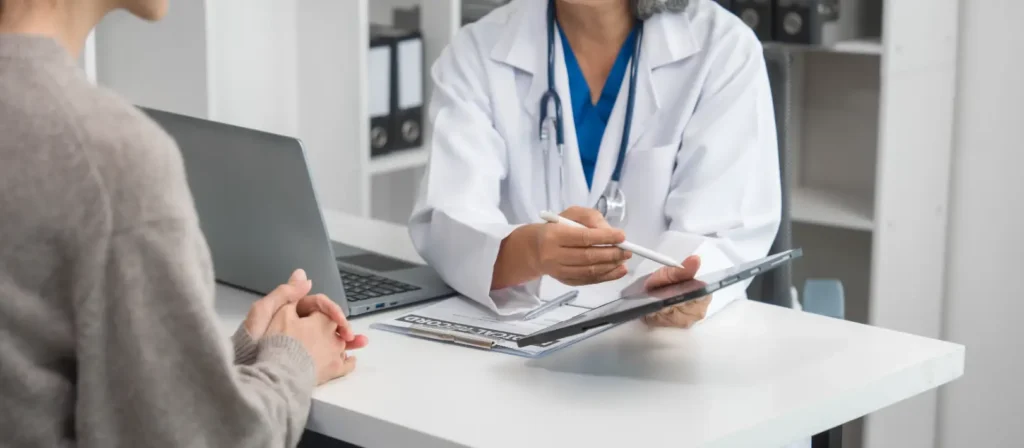 Doctor with a tablet speaks with a patient across a desk beside an open laptop and clipboard.