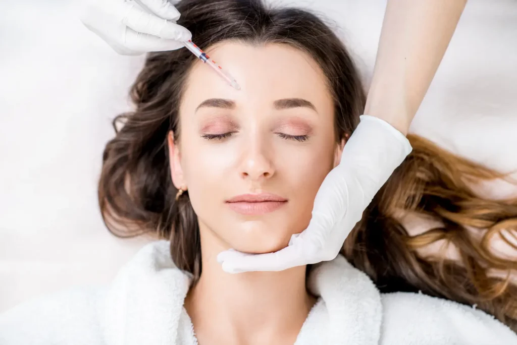 Woman lies with eyes closed while a gloved practitioner positions a syringe near her forehead.