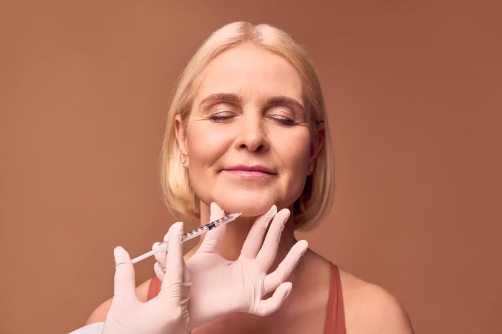 Mature woman with closed eyes as gloved hands position a syringe near her chin.