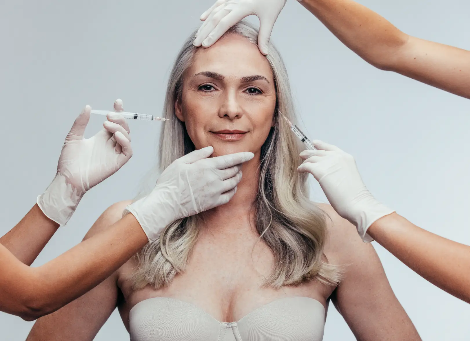 Mature woman surrounded by gloved hands holding syringes near her cheeks while another hand supports her chin.