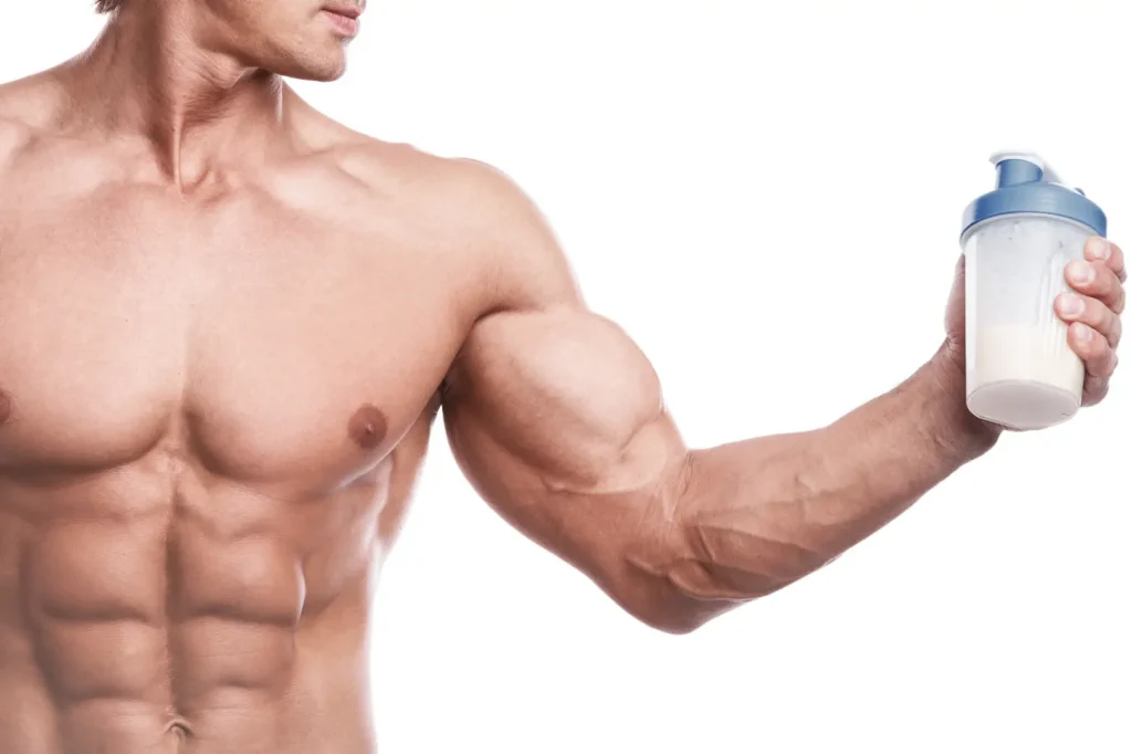 Shirtless muscular man holds a shaker bottle containing a light-colored drink against a white background.