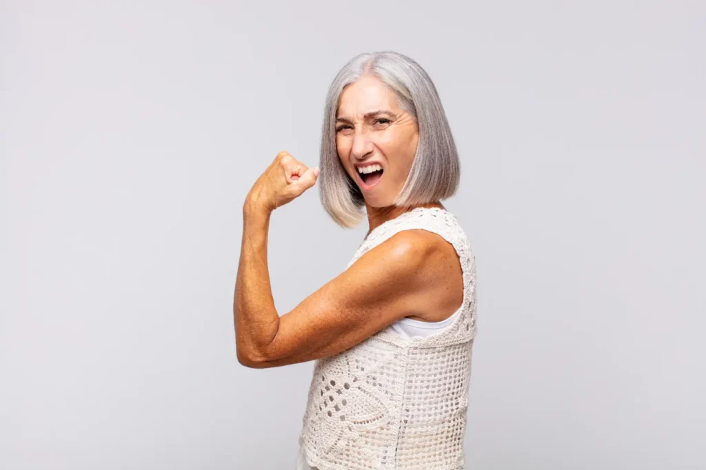 Older woman with gray hair flexes one arm while standing against a light gray studio background.