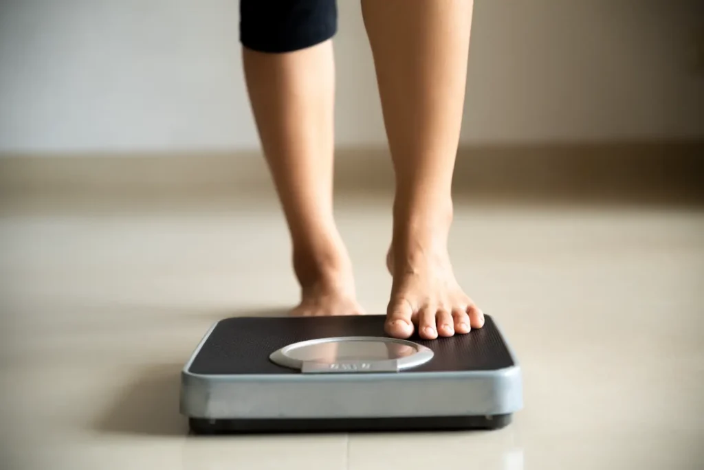 Barefoot person places one foot onto a bathroom scale positioned on a light-colored floor.