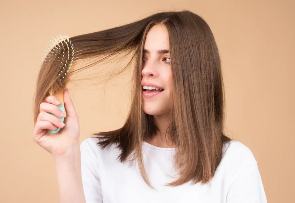 Woman in a white shirt holds a hairbrush through a section of her long brown hair.