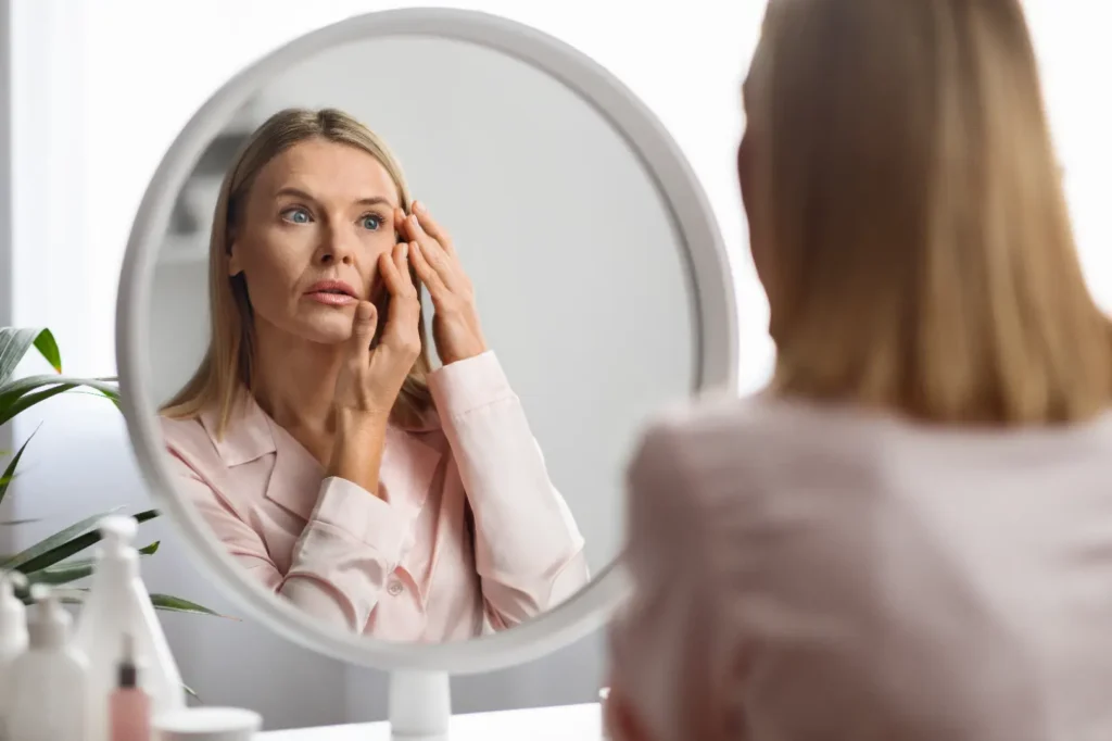 Woman looks into a round mirror while touching the skin beside her eye with both hands.