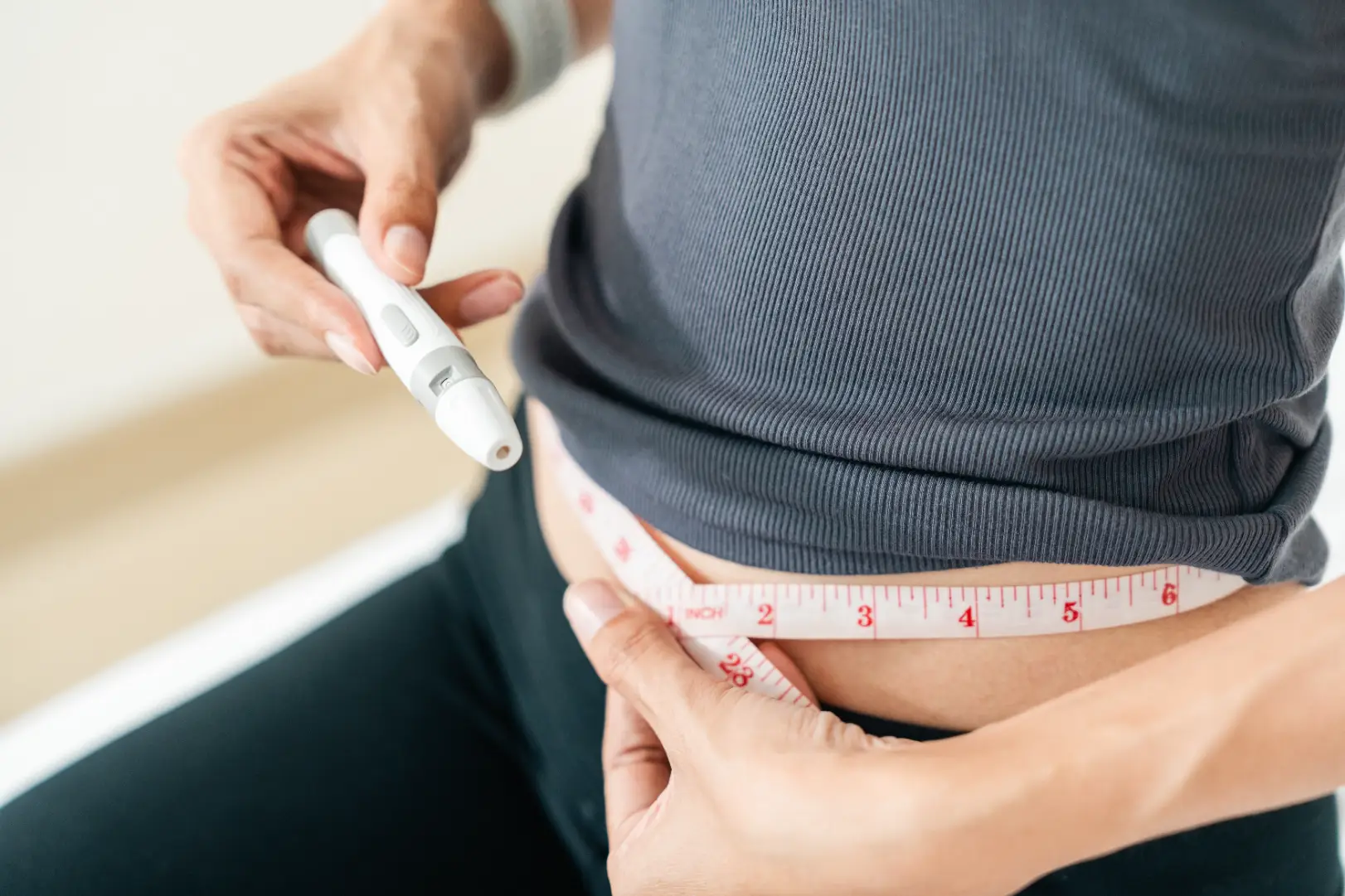 Woman wraps a measuring tape around her waist while holding a white injection pen near her abdomen.