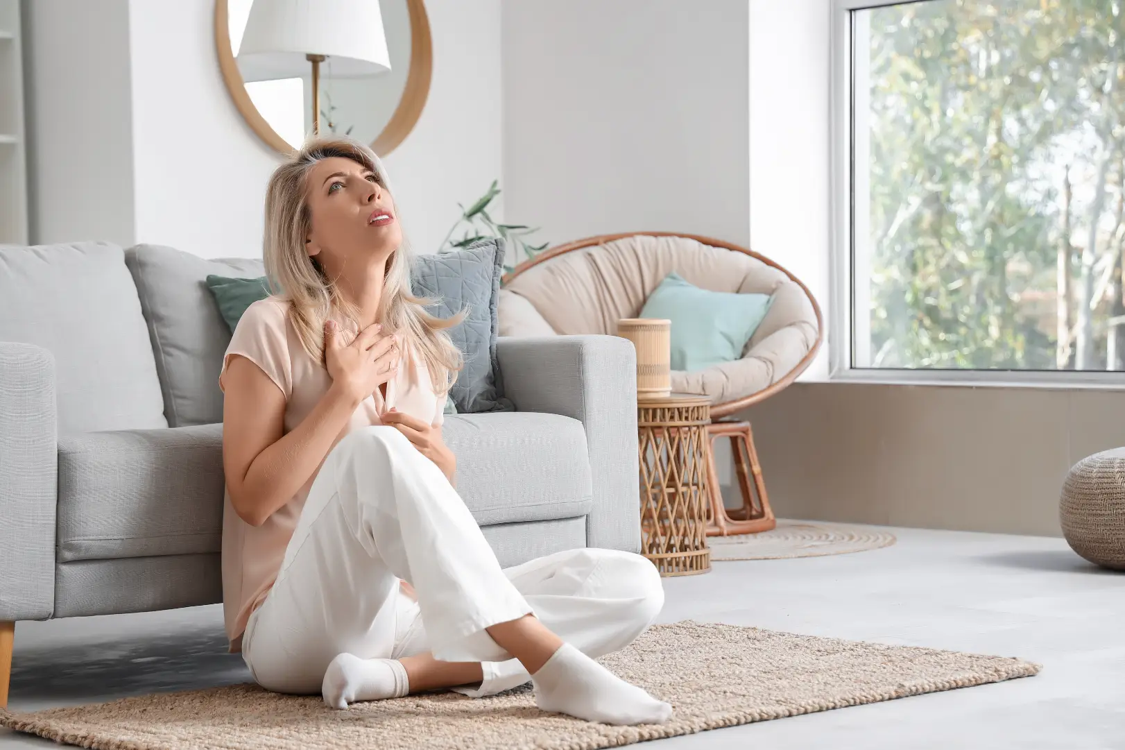 Woman sits on a rug beside a sofa with one hand on her upper chest and the other near her abdomen.